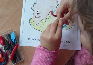 Przy stoliku siedzi dziewczynka w różowej bluzce w białe serca. Uczennica wykleja plasteliną aparat słuchowy widoczny na kolorowance. Na stoliku leżą przybory szkolne i plastelina.