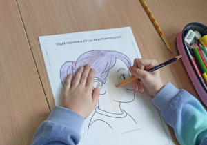 Uczennica w niebiesko-fioletowej bluzce koloruje rysunek przedstawiający dziewczynkę z aparatem słuchowym. Obok rysunku leży piórnik z przyborami szkolnymi.