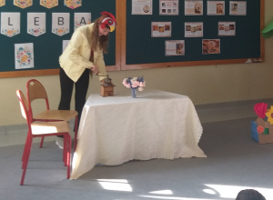 Na zdjęciu widać dziewczynkę stojącą przy stole, przykrytym obrusem. Uczennica ma na głowie czapkę koguta. Na stole stoi niewielki młynek ręczny, obok młynka wazonik z kwiatami.