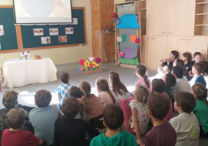 Na zdjęciu widać grupę dzieci siedzących na podłodze w świetlicy i patrzących w stronę scenki przygotowanej z przodu pomieszczenia. Przed nimi znajduje się stół przykryty białym obrusem, na którym stoi wazon z kwiatami i bochenek chleba. Obok stołu widać dekorację z papierowych kwiatów i domek z kartonu. Na ścianie wisi ekran, na którym wyświetlany jest obraz przedstawiający kamienny żarn ( mielenie ziarna). W tle widać też tablicę z przyczepionymi ilustracjami i tekstami o chlebie.