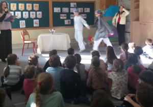 Na zdjęciu widać uczniów biorących udział w przedstawieniu. Uczennica– narrator, trzyma mikrofon, dwie dziewczynki, przebrane za myszki, tańczą. Po przeciwnej stronie stoi dziecko przebrane za koguta. Z tyłu stoi stół przykryty obrusem, wazon z kwiatami. Obok dekoracje z papierowych kwiatów i domek z kartonu. W tle na szkolnej tablicy widnieje napis : Dzień Chleba, oraz umieszone plakaty o chlebie.