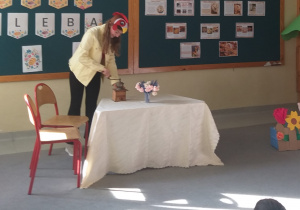 Na zdjęciu widać dziewczynkę stojącą przy stole, przykrytym obrusem. Uczennica ma na głowie czapkę koguta. Na stole stoi niewielki młynek ręczny, obok młynka wazonik z kwiatami. Dziecko odgrywające rolę, pokazuję jak wygląda proces mielenia ziarna. Z jednej strony stołu – dwa krzesła, z drugiej – wykonany z papieru płotek z kwiatami. W tle widać dużą tablicę z przyczepionymi plakatami o chlebie, oraz litery układające się w napis „ Dzień Chleba”.