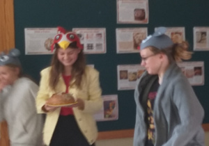 Na zdjęciu widać uczennicę przebraną za koguta, która trzyma na talerzu okrągły chlebek. Obok widać dwie dziewczynki, przebrane za myszki.