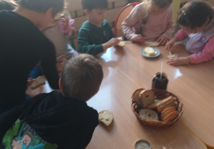 Na zdjęciu widać grupę dzieci siedzących wokół drewnianego stołu. Każde z nich ma kromkę chleba, którą smaruje masłem lub dżemem. Na środku stołu stoi koszyk z pokrojonym chlebem oraz słoik z dżemem.