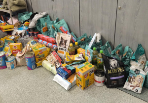 Na zdjęciu widać dużą ilość zebranych darów dla zwierząt, ustawionych na podłodze przy szafkach. Wśród nich znajdują się worki suchej karmy, puszki z mokrą karmą, smakołyki, makarony, oleje oraz inne produkty spożywcze. Widać różne marki karm, m.in. ActivPet, Pedigree, Butcher’s, czy Tropi. Z tyłu leżą również torby i koce, które mogą służyć jako legowiska dla zwierząt.