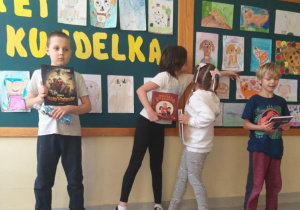 Na zdjęciu widać grupę czworga dzieci stojących przed dużą tablicą na ścianie. Tablica jest ozdobiona napisem z żółtych liter oraz wieloma kolorowymi rysunkami przedstawiającymi psy wykonanymi przez dzieci. Dzieci stoją w szeregu przed tablicą i trzymają w dłoniach książki lub zeszyty. Jedno z dzieci po lewej stronie trzyma książkę i butelkę wody. Dwoje dzieci pośrodku ogląda lub wskazuje rysunki na tablicy. Dziecko po prawej stronie również trzyma książkę lub zeszyt.