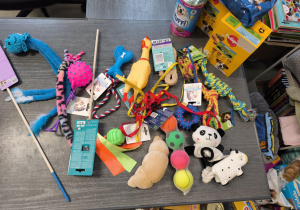 Na blacie stołu znajdują się m.in.:pluszowe zabawki (np. panda, kurczak-piszczałka, pluszowy rogalik), sznurki do przeciągania w różnych kolorach i grubościach, piłki – gumowe, tenisowe, z wypustkami, a także siatka z kilkoma piłkami, zabawki na sznurku oraz z piszczałkami.