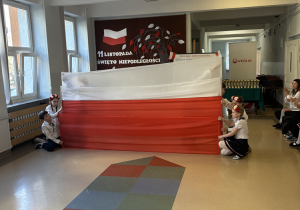 Na zdjęciu widać dzieci w szkolnym korytarzu podczas uroczystości z okazji Święta Niepodległości. Na środku trzymają dużą, poziomą flagę Polski w barwach biało-czerwonych, rozciągniętą szeroko na całą długość korytarza. W tle znajduje się dekoracja z napisem „11 listopada – Święto Niepodległości” oraz biało-czerwoną flagą i stylizowanym drzewem z liśćmi w barwach narodowych. Obok widoczny jest także baner z informacją o „XXX Biegu Niepodległości” oraz logo sponsora – Veolia. Dzieci ubrane są odświętnie: dziewczynki mają białe bluzki, ciemne spódniczki z biało-czerwonymi lamówkami i wianki z kwiatów w barwach narodowych, a chłopiec po lewej stronie ma białą koszulę i ciemne spodnie.