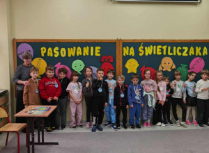 Na tle napisu 'Pasowanie na świetliczaka' stoją dzieci z zawieszonymi na szyi odznakami w kolorze niebieskim i różowym.