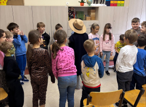 Dzieci stoją w ciasnym kręgu na środku klasy. W centrum stoi dziecko w dużym beżowym kapeluszu i ciemnym płaszczu, jakby odgrywało rolę w zabawie lub przedstawieniu.