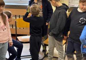 Dzieci stoją w kręgu. Dziecko w kapeluszu i płaszczu, z lupą w dłoni przygląda się chłopcu ubranemu na czarno, stojącemu tyłem do aparatu. Pozostali uczniowie przyglądają się tej scenie.