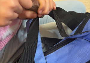 Zbliżenie na dłonie dziecka trzymającego pasek niebieskiej torby. Dziecko w różowym rękawie zaciska dłonie na czarnym pasku i drewnianym przedmiocie w kształcie walca. Tło jest rozmyte, a widać jedynie fragmenty innych dzieci siedzących na podłodze.