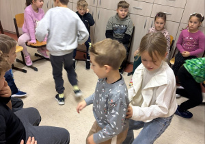 W sali lekcyjnej grupa dzieci bawi się w kręgu ustawionym z krzeseł. Kilkoro dzieci chodzi między krzesłami, trwa zabawa w „sałatkę owocową".  Pozostałe dzieci siedzą na krzesłach i obserwują. Wszyscy wyglądają na zaangażowanych i uśmiechniętych. Tło stanowią jasne szafki, a na nich pudełko, globus i kilka dekoracji.