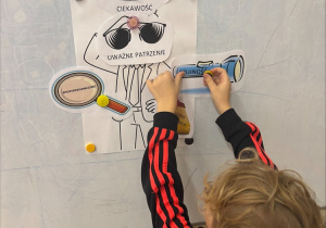 Dziecko stoi przy białej tablicy i przypina magnesami papierowe elementy przedstawiające detektywa. Na rysunku są podpisy po polsku, m.in. „Ciekawość”, „Uważne patrzenie” i „Spostrzegawczość”. Dziecko ma jasne, kręcone włosy i nosi czarną bluzę z czerwonymi paskami na rękawach. Wygląda to na zadanie związane z rozwijaniem umiejętności detektywistycznych lub spostrzegawczości.