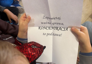 Dziecko trzyma białą kartkę z napisem " Zapamiętaj, ważna sprawa, koncentracja to podstawa!"