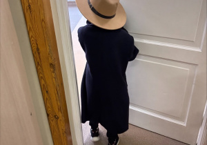 Dziecko ubrane w ciemny płaszcz i duży, beżowy kapelusz stoi tyłem do aparatu i otwiera drzwi klasy. Wygląda, jakby wychodziło na korytarz. Korytarz jest jasno oświetlony, a podłoga w klasie ma ciemnobrązowy odcień. Obraz sprawia wrażenie sceny z zabawy lub małego przedstawienia.