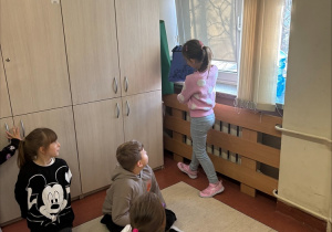 Kilkoro dzieci znajduje się w sali szkolnej. Troje z nich klęczy na podłodze i patrzy w stronę dziewczynki stojącej przy oknie. Dziewczynka stoi tyłem, ubrana w różowy sweter i jasnoniebieskie spodnie, i zagląda do niebieskiej torby postawionej na małym stoliku. W pomieszczeniu widać jasne szafki i duże okno z opuszczonymi roletami. Scena wygląda na część zajęć szkolnych lub zabawy.