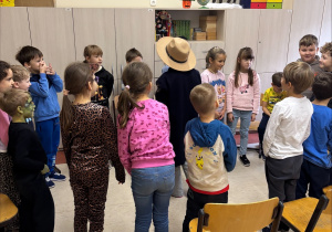 Dzieci stoją w ciasnym kręgu na środku klasy. W centrum stoi dziecko w dużym beżowym kapeluszu i ciemnym płaszczu, jakby odgrywało rolę w zabawie lub przedstawieniu. Pozostałe dzieci patrzą na nie z zainteresowaniem. W tle znajdują się drewniane krzesła oraz jasne szafki, na których widać globus, ozdobę w kształcie renifera i kolorowe pudełka.