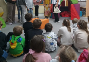 Na zdjęciu widać dzieci siedzące na podłodze i oglądające przedstawienia. Grupa dzieci wciela się w role bohaterów inscenizacji. Występujący uczestnicy mają na głowach kolorowe maski, przedstawiające różne zwierzątka. Po prawej stronie – dziewczynka trzyma mikrofon, a po lewej – dziewczynka grająca rolę czapli. W głębi sceny widoczni są pozostali uczestnicy przedstawienia. Na podłodze ustawiono pufy, symbolizujące domki zwierząt. Z tyłu na tablicy umieszczona dekoracja: papierowe promienne słońce oraz drzewo z liśćmi zawierającymi hasła do okazywania życzliwości, a także fragment napisu „Życzliwość, podaj dalej”.