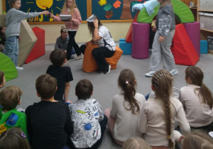 Zdjęcie przedstawia dzieci wcielające się w role bohaterów inscenizacji. Uczniowie noszą na głowach maski zwierząt. Po prawej stronie dziewczynka gra czaplę, pośrodku – siedząca na pufie uczennica odgrywa rolę borsuka. Z lewej stoi chłopiec – zajączek, obok którego znajduje się narratorka, a za nią siedzi kolejny uczestnik przedstawienia. Na podłodze ustawiono pufy przedstawiające domki zwierząt. Przed sceną na podłodze siedzą dzieci stanowiące publiczność. W tle widoczna jest dekoracja: papierowe promienne słońce oraz drzewo z liśćmi zawierającymi hasła do okazywania życzliwości, a także fragment napisu „Życzliwość, podaj dalej”.