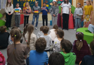 Na zdjęciu uchwycono moment, w którym występujący uczniowie prezentują flagi państw wykonane z papieru. Na podłodze siedzi publiczność – dzieci oglądają przedstawienie. W tle znajduje się dekoracja na tablicy: uśmiechnięte papierowe słońce oraz drzewo z liśćmi zawierającymi hasła zachęcające do okazywania życzliwości. W tle widać również fragment napisu „Życzliwość, podaj dalej”.