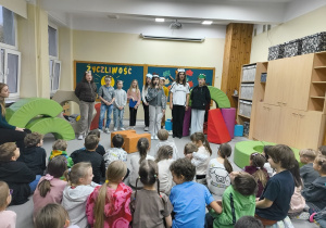 Na zdjęciu widać dzieci siedzące na podłodze i oglądające przedstawienia. Występujący uczestnicy mają na głowach kolorowe maski, przedstawiające różne zwierzątka. Na środku stoi dziewczynka i trzyma mikrofon. Za nią pozostali uczestnicy. . Na podłodze ustawiono pufy, symbolizujące domki zwierząt. Z tyłu na tablicy umieszczona dekoracja: papierowe promienne słońce oraz drzewo z liśćmi zawierającymi hasła do okazywania życzliwości, a także fragment napisu „Życzliwość, podaj dalej”.