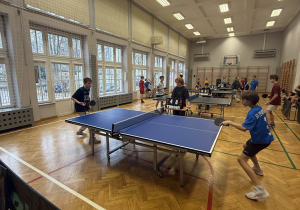 Na zdjęciu widać mecz tenisa stołowego rozgrywany w szkolnej hali sportowej. Dwóch chłopców gra przy stole na pierwszym planie — jeden po lewej stronie w ciemnej koszulce, drugi po prawej w niebieskiej koszulce. Wyglądają na mocno skoncentrowanych i zaangażowanych w wymianę.