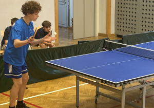 Zawodnik stoi przy stole pingpongowym i przygotowuje się do wykonania serwu – w lewej ręce unosi piłeczkę, a w prawej trzyma rakietkę. Ma na sobie sportowy strój: niebieską koszulkę i krótkie spodenki, a także sportowe buty. W tle widać inne dzieci siedzące na krzesłach i obserwujące rozgrywkę. Sala ma drewnianą podłogę, a stół do tenisa stołowego jest profesjonalny, z siatką i boczną osłoną.