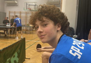 Na zdjęciu widać młodego chłopaka siedzącego na ławce przy stole do tenisa stołowego w hali sportowej. Ma kręcone, brązowe włosy i nosi niebieską sportową koszulkę z białym nadrukiem na plecach. W ręku trzyma rakietkę pingpongową, a obok niego siedzą inni zawodnicy. W tle widać rozgrywkę przy stole tenisowym oraz kilka osób obserwujących zawody.
