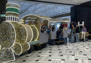 Na zdjęciu widać grupę dzieci oraz kilka osób dorosłych pozujących przy dużej, dekoracyjnej instalacji w kształcie świątecznego pociągu. Lokomotywa i wagony są bogato ozdobione złotymi, błyszczącymi elementami oraz podświetlane jasnymi lampkami, co tworzy efektowną, świąteczną atmosferę. Część dzieci siedzi lub stoi w wagonikach, wyglądając przez okna i uśmiechając się do zdjęcia, inne stoją obok pociągu. Cała scena znajduje się w przestrzeni centrum handlowego – widać jasną, nowoczesną posadzkę oraz szklany dach w tle.