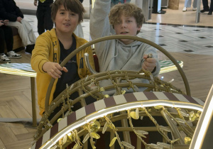 Na zdjęciu widać dwóch chłopców siedzących w ozdobnej, świątecznej instalacji przypominającej lokomotywę lub wagonik pociągu. Trzymają dużą dekoracyjną kierownicę i wyglądają na zaangażowanych w zabawę — jeden z nich podnosi rękę, jakby machał lub udawał sygnał maszynisty. Dekoracja jest bogato zdobiona złotymi elementami i podświetlana jasnymi lampkami, co dodaje jej świątecznego blasku. W tle widać wnętrze centrum handlowego oraz inne dzieci i dorosłych, którzy także biorą udział w mikołajkowym wyjściu.