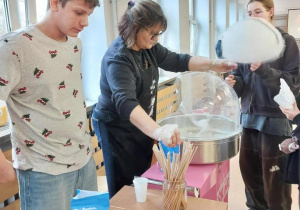 Na zdjęciu widać przygotowywanie waty cukrowej. W centrum znajduje się maszyna do waty cukrowej, przy której stoi dorosła osoba obsługująca urządzenie i formująca watę na patyku. Obok stoją młodzi ludzie, obserwujący proces i czekający na swoją porcję. Na stole przed maszyną leżą drewniane patyczki, opakowanie cukru oraz jednorazowy kubek. W tle widać duże okna, przez które wpada naturalne światło, oraz fragmenty szkolnego wyposażenia.