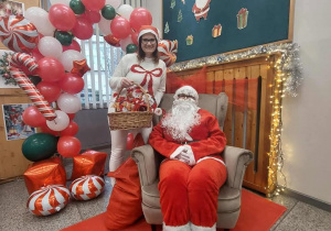 Po lewej stronie stoi ubrana choinka z lampkami i bombkami, a pod nią znajdują się worki i prezenty. Obok choinki ustawiono dekoracyjną bramę z balonów w kolorach czerwonym, białym i zielonym, ozdobioną motywami cukrowych lasek. Po prawej stronie na fotelu siedzi Mikołaj – w czerwonym stroju, z białą brodą i czarnymi butami. Za nim znajduje się dekoracja przypominająca kominek, ozdobiona światełkami i girlandą. Nad „kominkiem” wisi tablica z napisem „Mikołajki” oraz świątecznymi rysunkami, takimi jak choinki, prezenty i Mikołaj.