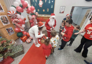 W centrum stoi Mikołaj w czerwonym stroju, a obok niego osoba pomagająca rozdawać prezenty. Przed nimi ustawione są dzieci, które podchodzą po upominki.Tło jest bogato udekorowane: łuk z balonów w kolorach czerwonym, białym i zielonym, ozdoby w kształcie lasek cukrowych, choinek i prezentów oraz świąteczna tablica z motywami bożonarodzeniowymi. Na podłodze leży czerwony dywan, a obok widać zapakowane prezenty.