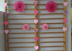 Na zdjęciu widać fragment szkolnego korytarza z drewnianą drabinką gimnastyczną przymocowaną do ściany. Drabinka została udekorowana w walentynkowym stylu. Zawieszono na niej różowe, papierowe rozety w różnych rozmiarach – jedną dużą u góry oraz kilka mniejszych poniżej. Po obu stronach drabinki oraz wzdłuż jej środka rozmieszczono papierowe serduszka w odcieniach różu i czerwieni. Serduszka tworzą pionowe girlandy, które nadają dekoracji symetrii i lekkości.
