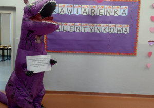 W centrum uwagi znajduje się osoba przebrana za dużego, fioletowego dinozaura w dmuchanym kostiumie. Dinozaur trzyma w łapkach kartkę z napisem: „Dinozaury nie jadły ciasteczek w naszej kawiarence… wyginęły. Zapraszamy do kawiarenki walentynkowej!” Hasło ma żartobliwy charakter i zachęca do odwiedzenia kawiarenki. W tle widoczna jest duża tablica ozdobiona fioletowym papierem z napisem „Kawiarenka Walentynkowa”. Litery umieszczone są na białych kartkach z walentynkowymi motywami. Obramowanie tablicy stanowią małe czerwone serduszka, a po prawej stronie ściany przyklejono dodatkowe różowe serca, które dopełniają dekorację.