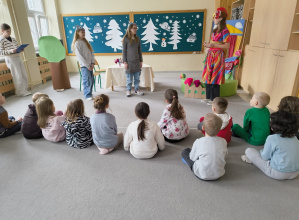 3.Na zdjęciu widać dzieci oglądające szkolne przedstawienie. Na pierwszym planie stoi uczennica pełniąca rolę narratora – w ręku trzyma tekst i z zaangażowaniem odczytuje swoją kwestię.