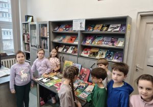 Na zdjęciu widać grupę dzieci stojących w bibliotece. Kilkoro uczniów stoi przy stoliku, na którym rozłożone są różne książki o kotach. Dzieci oglądają książki i rozmawiają między sobą. Niektóre patrzą na książki, a inne spoglądają w stronę aparatu. Za dziećmi znajdują się regały z książkami. Na półkach ustawiono wiele kolorowych książek, również o tematyce kotów. Na środku regału znajduje się kartka z napisem „Światowy Dzień Kota”, co sugeruje, że wystawa książek została przygotowana z okazji tego święta.