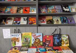 Na zdjęciu widać regał z książkami w bibliotece, na którym przygotowano specjalną wystawkę poświęconą kotom. Na górnej półce znajduje się plakat z napisem „ŚWIATOWY DZIEŃ KOTA” oraz zdjęciem kota. Pod plakatem ustawione są różne książki o kotach – zarówno opowiadania, jak i książki popularnonaukowe. Okładki są kolorowe i przedstawiają koty w różnych stylach – realistycznych i rysunkowych. Na środkowych półkach stoją kolejne książki o podobnej tematyce. Wśród nich można zauważyć tytuły dotyczące przygód kotów, ich życia, a także historie z udziałem innych zwierząt, np. tygrysów czy lwów. Przed regałem ustawiono stół, na którym wyłożono jeszcze więcej książek. Są one ułożone w kilku rzędach tak, aby było dobrze widać ich okładki. Wiele z nich przedstawia kocięta, dorosłe koty lub bohaterów – koty z bajek i opowiadań.