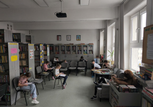 Na zdjęciu widać jasną, przestronną szkolną bibliotekę lub czytelnię. W pomieszczeniu znajduje się kilka rzędów szarych krzeseł i stolików, przy których siedzą dzieci. Większość z nich czyta książki lub przegląda materiały. Dzieci siedzą w różnych miejscach – przy stolikach, na krzesłach ustawionych w rzędach, a jedno z nich siedzi także w wygodnym fotelu po prawej stronie. Wzdłuż ścian stoją wysokie regały z książkami, wypełnione wieloma kolorowymi publikacjami. Na niektórych półkach znajdują się kartki i plakaty z informacjami lub pracami uczniów. Na ścianie wiszą też ramki z rysunkami. Po prawej stronie pomieszczenia znajdują się duże okna, przez które wpada dużo naturalnego światła. Na parapecie stoi roślina doniczkowa, a obok znajduje się stolik z książkami oraz duży pluszowy miś.