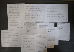 Na zdjęciu widać tablicę, na której przypięto kilka kartek z zeszytów i wydruków. Na kartkach znajdują się ręcznie napisane w języku polskim wiersze i krótkie teksty o kotach. Większość prac jest napisana przez dzieci – widać różne style pisma, podpisy z imionami oraz informację o klasie.