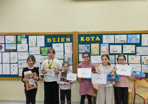 Zdjęcie przedstawia uroczyste wręczenia nagród z okazji Dni Kota. Na zdjęciu widać sześcioro dzieci stojących w szkolnej świetlicy, na tle gazetki ściennej z napisem „DZIEŃ KOTA”. Za nimi znajdują się dwie duże, zielone tablice w drewnianych ramach, wypełnione rysunkami kotów wykonanymi przez uczniów.