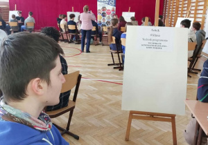 Na zdjęciu widać salę gimnastyczną lub dużą salę szkolną z drewnianą podłogą i drabinkami przy ścianach. Na końcu sali znajduje się duża, czerwona kurtyna. Przed nią ustawiono krzesła, na których siedzą uczniowie zwróceni w stronę przodu, jak podczas prezentacji lub konkursu. Na pierwszym planie po lewej stronie siedzi chłopiec w niebieskiej bluzie z kapturem i chustą pod szyją. Patrzy w stronę przodu sali. Obok niego stoi sztaluga z białą planszą. Na planszy widnieje napis z imieniem „Antek”, informacją „4 klasa” oraz „Technik programista – technikum kinematograficzno-komputerowe”. Z przodu sali stoi kobieta. W tle widać również stojak z plakatem informacyjnym dotyczącym szkoły.