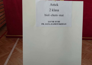 Na zdjęciu widać białą tablicę ustawioną na drewnianej sztaludze w sali gimnastycznej. W tle znajduje się duża czerwona kurtyna, a na podłodze widać drewniany parkiet z liniami do gier sportowych.