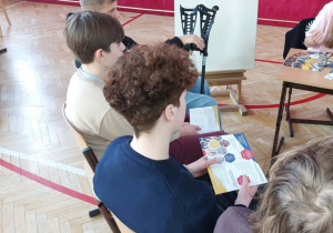 Na zdjęciu widać grupę dzieci siedzących na krzesłach w sali gimnastycznej lub szkolnej. Podłoga jest drewniana, z zaznaczonymi czerwonymi liniami do gier sportowych. W tle znajduje się duża czerwona kurtyna. Dzieci siedzą w rzędzie i są zwrócone w stronę tablicy ustawionej na sztaludze. Na tablicy znajduje się kartka z napisem, m.in. imię Natalia, informacja „4 klasa” oraz tekst związany z konkursem lub mistrzostwami szkolnymi. Obok tablicy stoi ozdobne czarne krzesło.