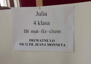 Na zdjęciu widać białą tablicę ustawioną na drewnianej sztaludze w sali gimnastycznej. W tle znajduje się duża czerwona kurtyna.