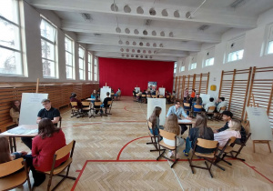 Na zdjęciu widać dużą szkolną salę gimnastyczną. Pomieszczenie jest jasne, z wysokim sufitem i dużymi oknami po lewej stronie, przez które wpada dużo światła dziennego. Podłoga jest drewniana z czerwonymi liniami do gier sportowych. Po prawej stronie znajdują się drabinki gimnastyczne. W sali ustawiono kilka stolików, przy których siedzą grupy uczniów. Każda grupa składa się z kilku osób siedzących na krzesłach i rozmawiających ze sobą lub wykonujących jakieś zadania. Przy niektórych stolikach stoją tablice na sztalugach z kartkami zawierającymi informacje.