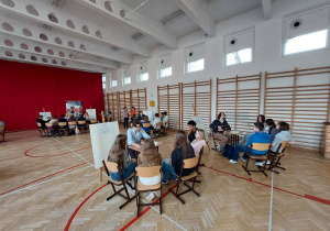 Na zdjęciu widać dużą szkolną salę gimnastyczną. Pomieszczenie jest jasne, z wysokim sufitem i dużymi oknami po lewej stronie, przez które wpada dużo światła dziennego. Podłoga jest drewniana z czerwonymi liniami do gier sportowych. Po prawej stronie znajdują się drabinki gimnastyczne. W sali ustawiono kilka stolików, przy których siedzą grupy uczniów. Każda grupa składa się z kilku osób siedzących na krzesłach i rozmawiających ze sobą lub wykonujących jakieś zadania. Przy niektórych stolikach stoją tablice na sztalugach z kartkami zawierającymi informacje.