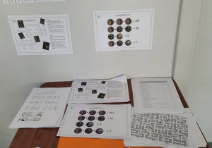 Na zdjęciu widać przygotowaną szkolną wystawkę z okazji Dnia Liczby π. Na białej ścianie nad stołem wiszą cztery plakaty z zadaniami matematycznymi. Jeden z nich ma tytuł „Dzień Liczby π” i zawiera informacje o tej liczbie oraz jej rozwinięciu dziesiętnym. Obok znajdują się różne łamigłówki, m.in. wykreślanka z nazwiskami znanych matematyków oraz zadanie obrazkowe z działaniami.