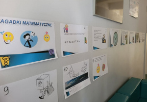 Na zdjęciu widać fragment szkolnego korytarza lub sali, na ścianie której rozwieszono kartki z zadaniami i zagadkami matematycznymi. U góry znajduje się większy plakat z napisem „ZAGADKI MATEMATYCZNE”, ozdobiony rysunkami i emotikonami sugerującymi myślenie i rozwiązywanie problemów.